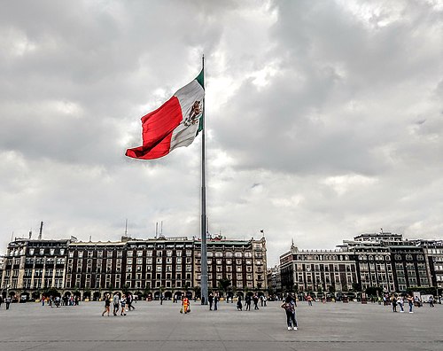 View of the Zócalo, also known as Plaza de la Constitución, in Mexico City, with the Mexican flag waving in the center and the Old Portal de Mercaderes to the right